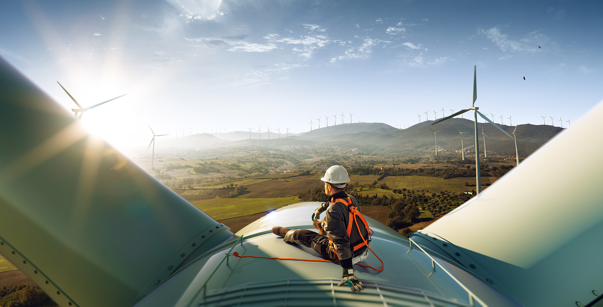 Un ingénieur est assis au soleil sur une éolienne, le regard tourné au-delà de champs verts en direction d'un parc éolien